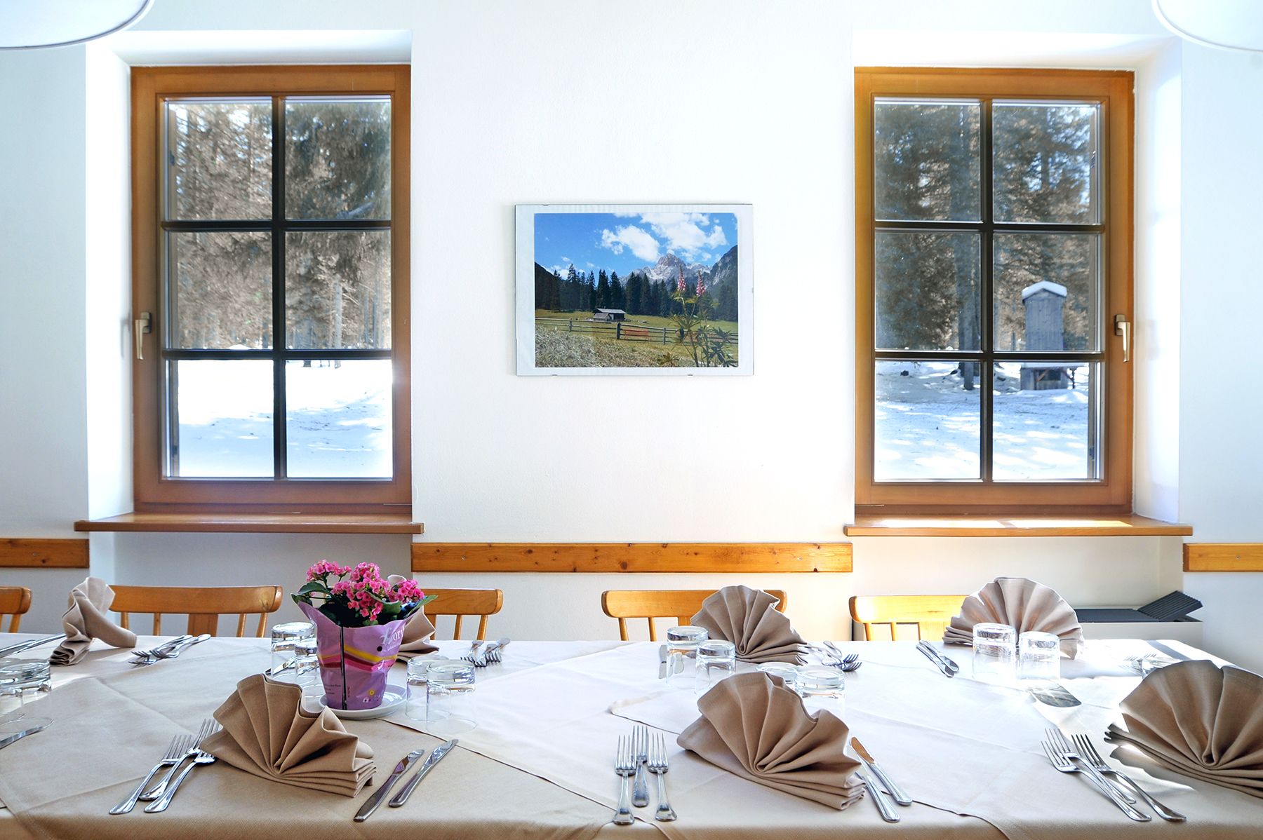 A dining room with a table set for four, two windows with a view of a snowy landscape, and a framed picture on the wall.