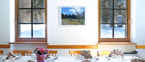 A dining room with a table set for four, two windows with a view of a snowy landscape, and a framed picture on the wall.