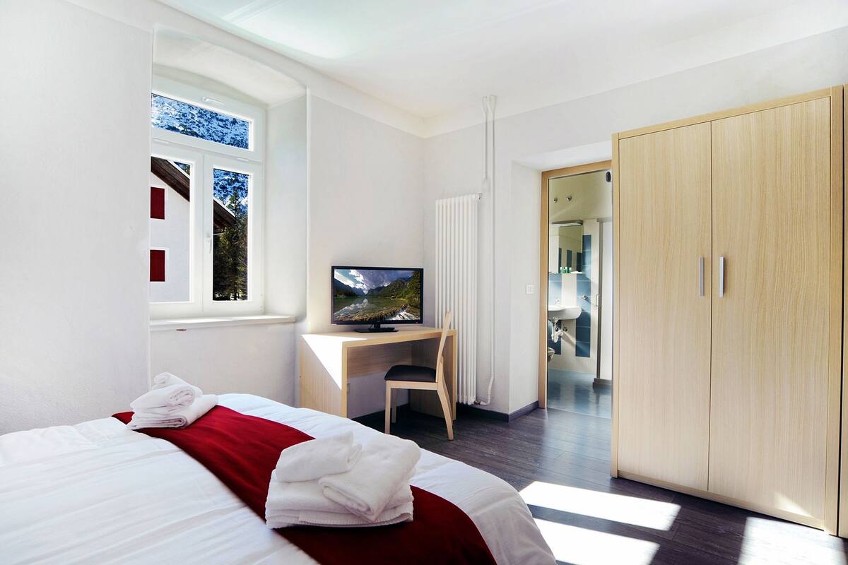A modern hotel room with a bed, a desk with a TV, a wardrobe, and a window with a view of trees.