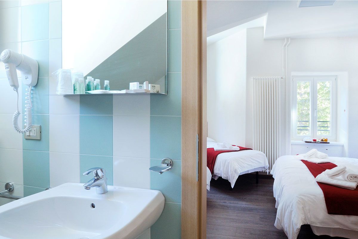 A hotel room with a bathroom, two beds, a window, and a radiator.