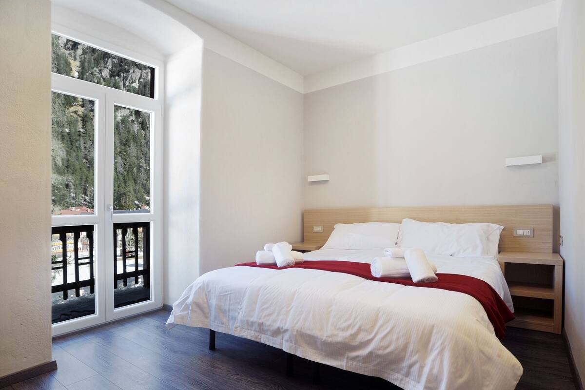 A hotel room with a large bed, a wooden headboard, a nightstand, and a window with a view of trees.