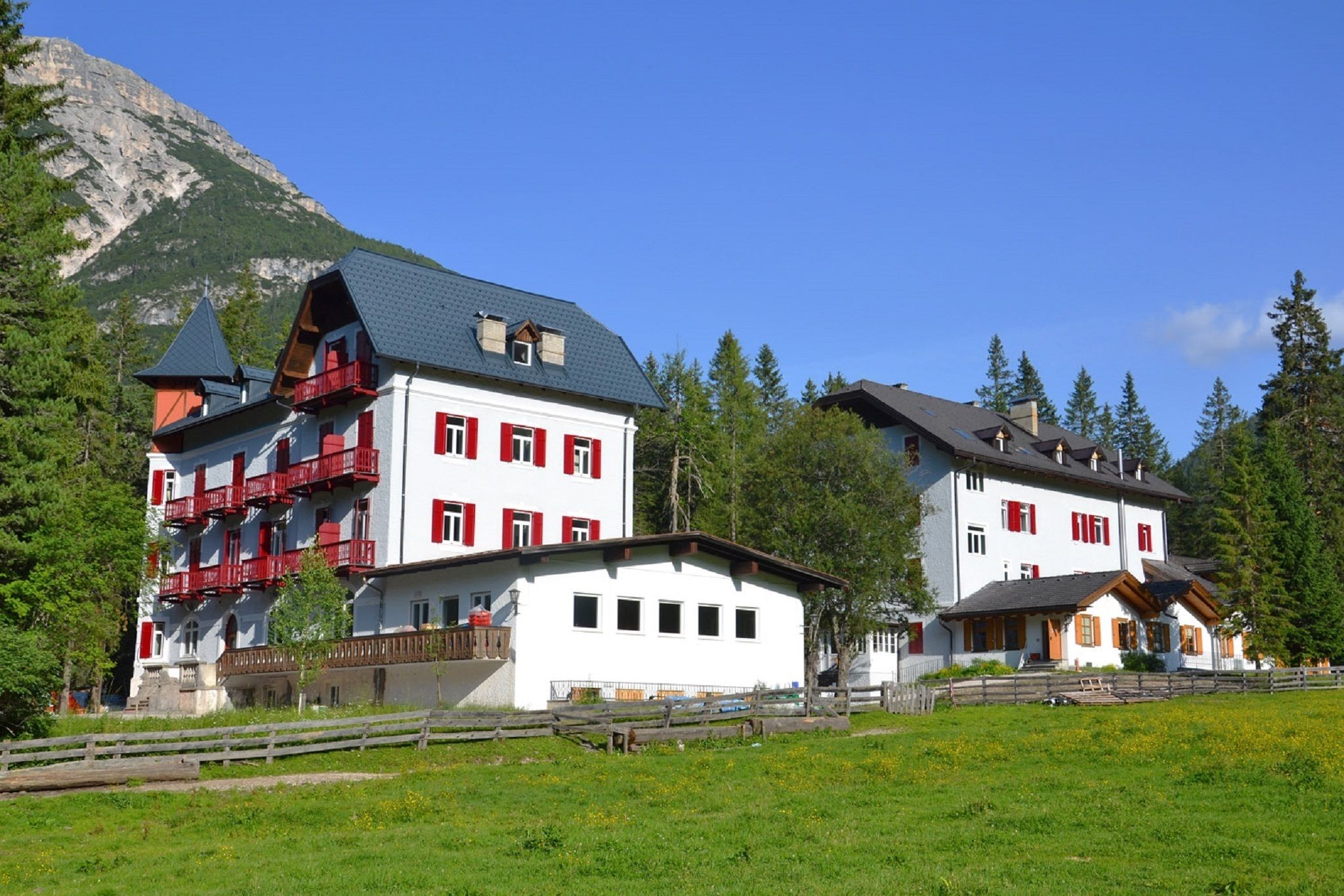 Un edificio grande de varios pisos con balcones rojos y paredes blancas, rodeado de un paisaje verde y montañas.