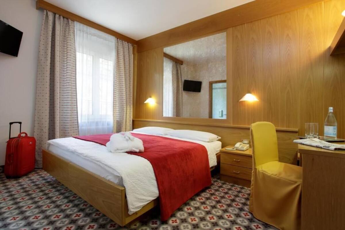 Habitación de hotel con cama, escritorio, silla, maleta y espejo.