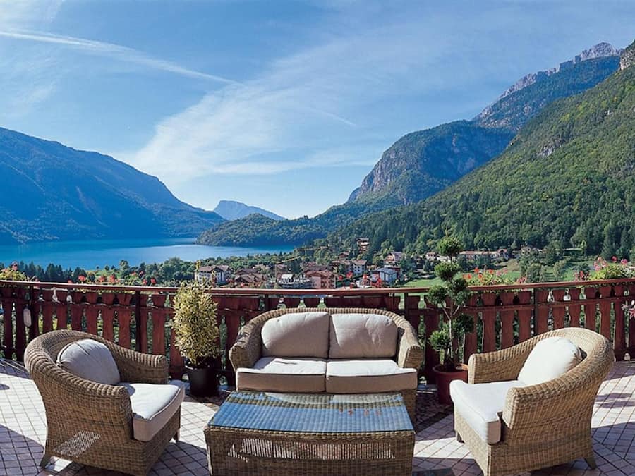 Um terraço com móveis de vime, vista para um lago e montanhas ao fundo.