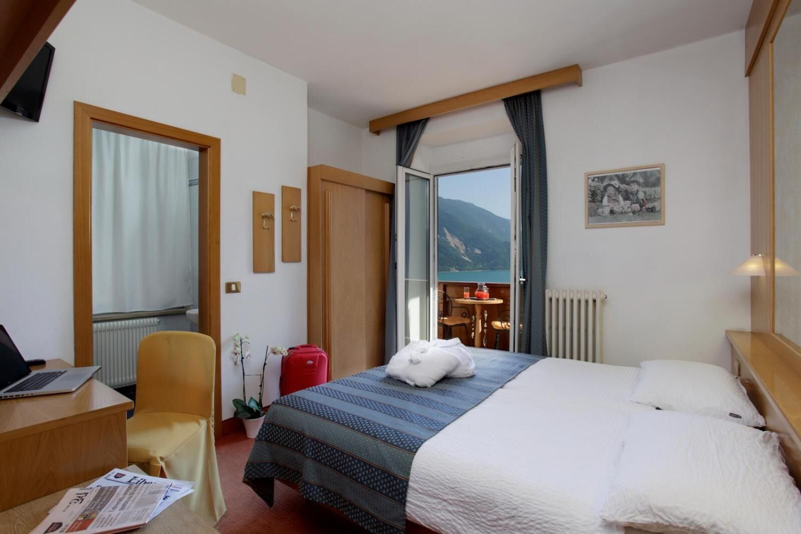 A hotel room with a bed, a desk, a chair, a red suitcase, and a view of mountains through an open door.