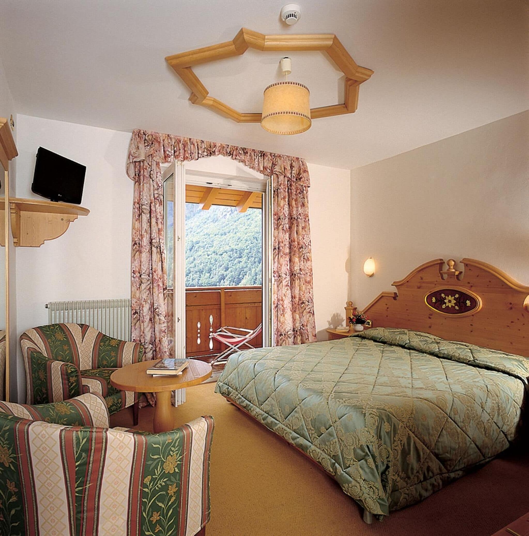 Quarto com cama, cadeira, mesa pequena, televisão em prateleira e janela com cortinas.
