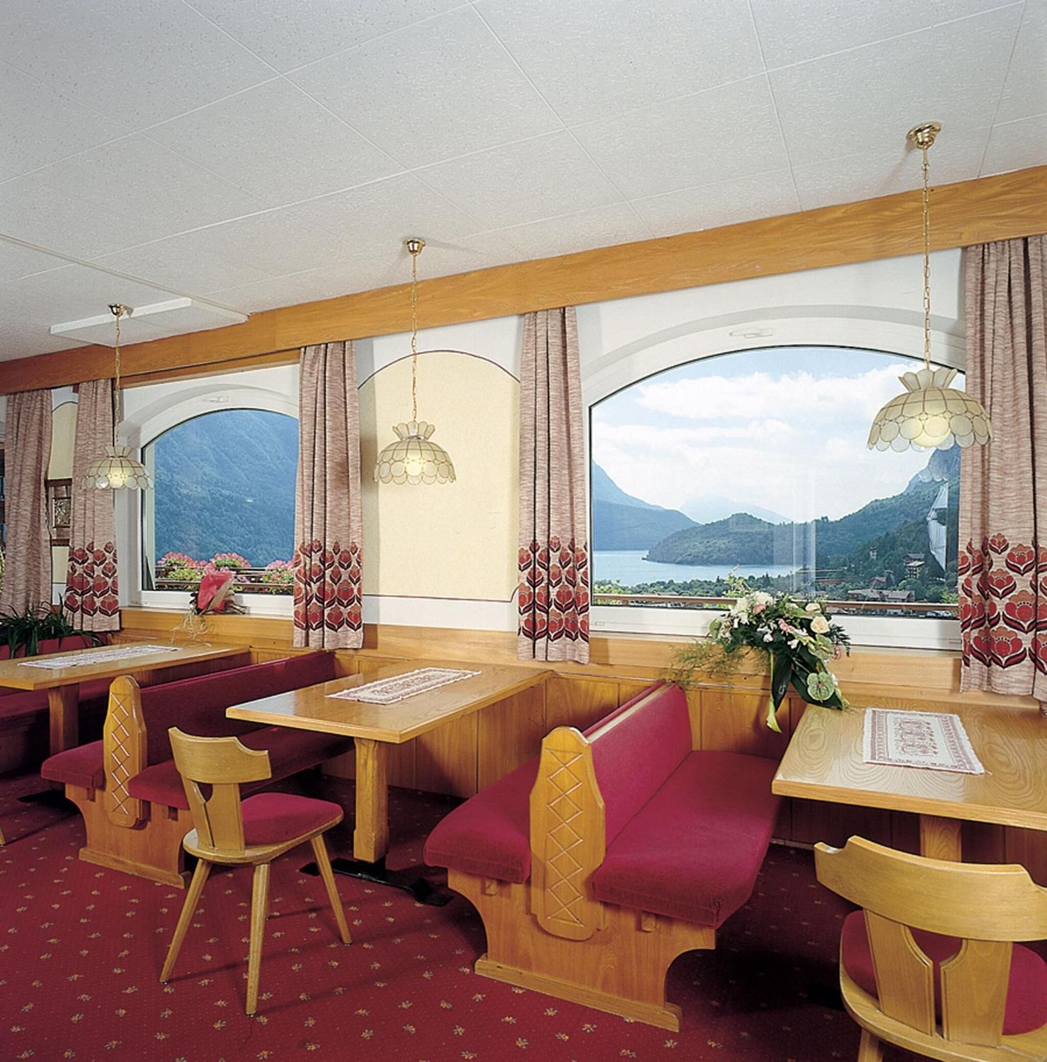 Un restaurante con mesas y bancos de madera, ventanas amplias con cortinas y lámparas colgantes.