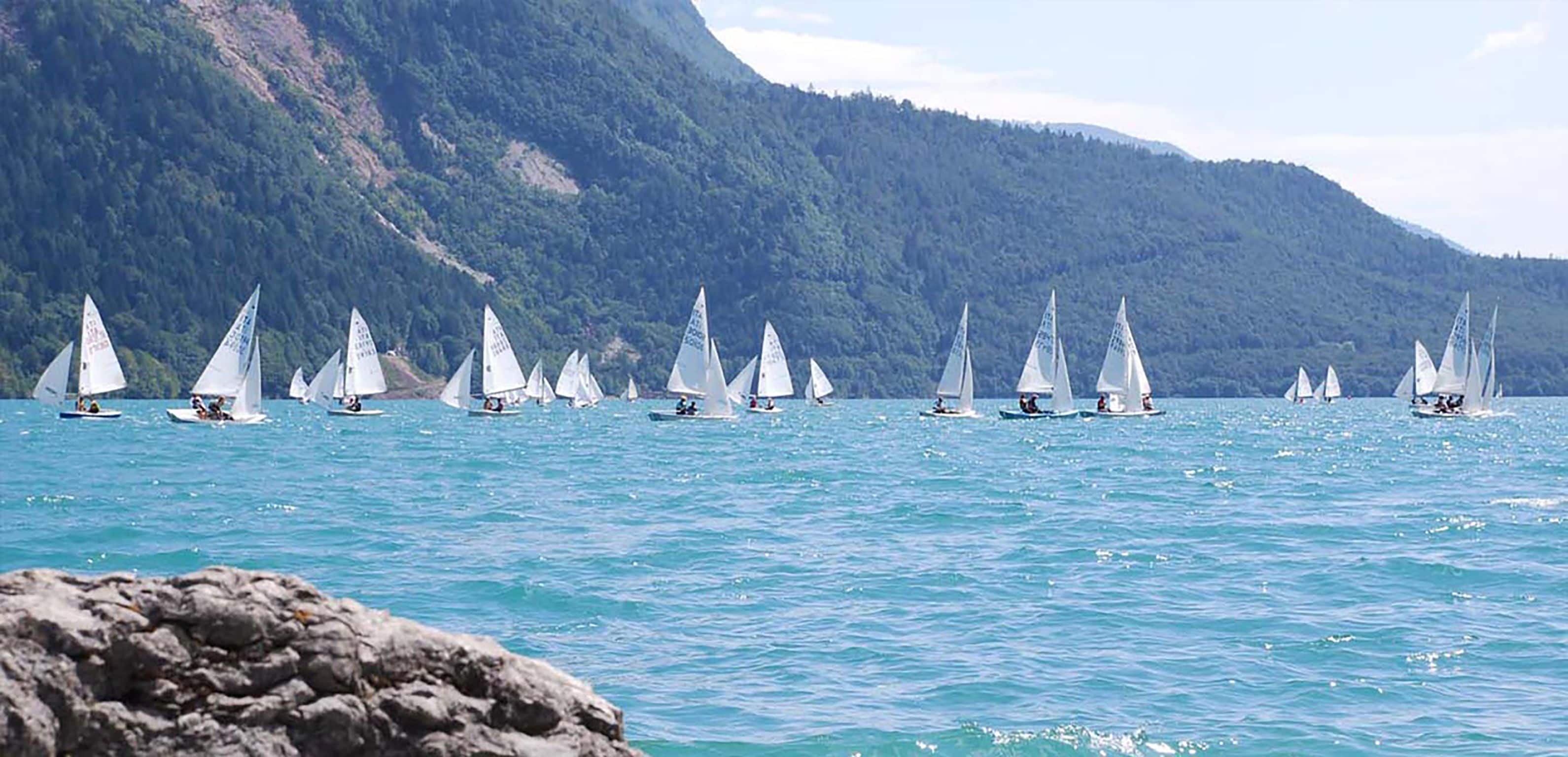 Eine Gruppe Segelboote auf ruhiger See, mit Bergen im Hintergrund.