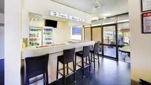 A bar area with a sign reading "The Rainwater Bar Welcome You All", bar stools, a refrigerator stocked with beverages, and a view of the outside through large windows. - Raintree Motel (Hermit Park)