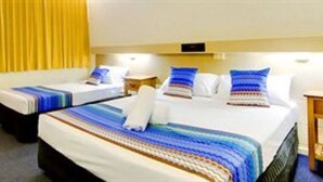 A hotel room with two single beds, each with a striped blanket and pillows, a wooden nightstand, and a desk with a chair. - Raintree Motel (Hermit Park)