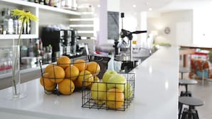 Una zona de bar con una barra que exhibe naranjas y manzanas, un florero con una flor amarilla y estantes llenos de botellas.