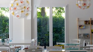 Un comedor luminoso y moderno con mobiliario blanco y sillas coloridas, ventanas amplias con vistas a la vegetación y lámparas decorativas colgantes.