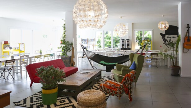 Amplia sala de estar con un sofá rojo, un sillón verde y una hamaca. Hay luces colgantes y un ventanal grande que proporciona luz natural.