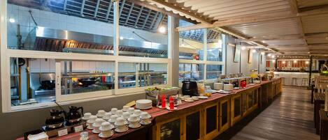 A long buffet table with various dishes and utensils, large windows, and a high ceiling with wooden beams.