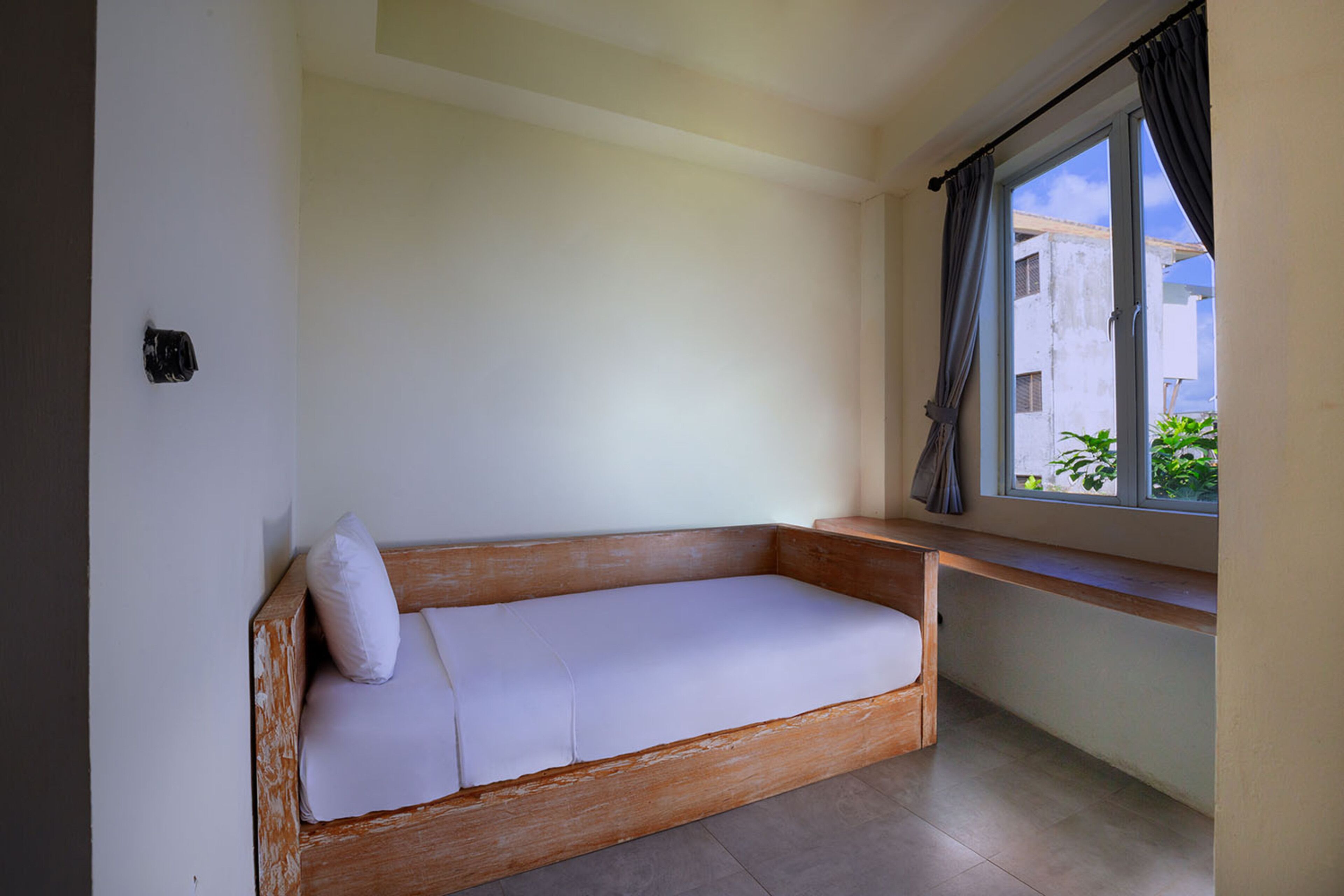 A small, single-room with a wooden bench bed, a window with curtains, and a view of buildings outside.