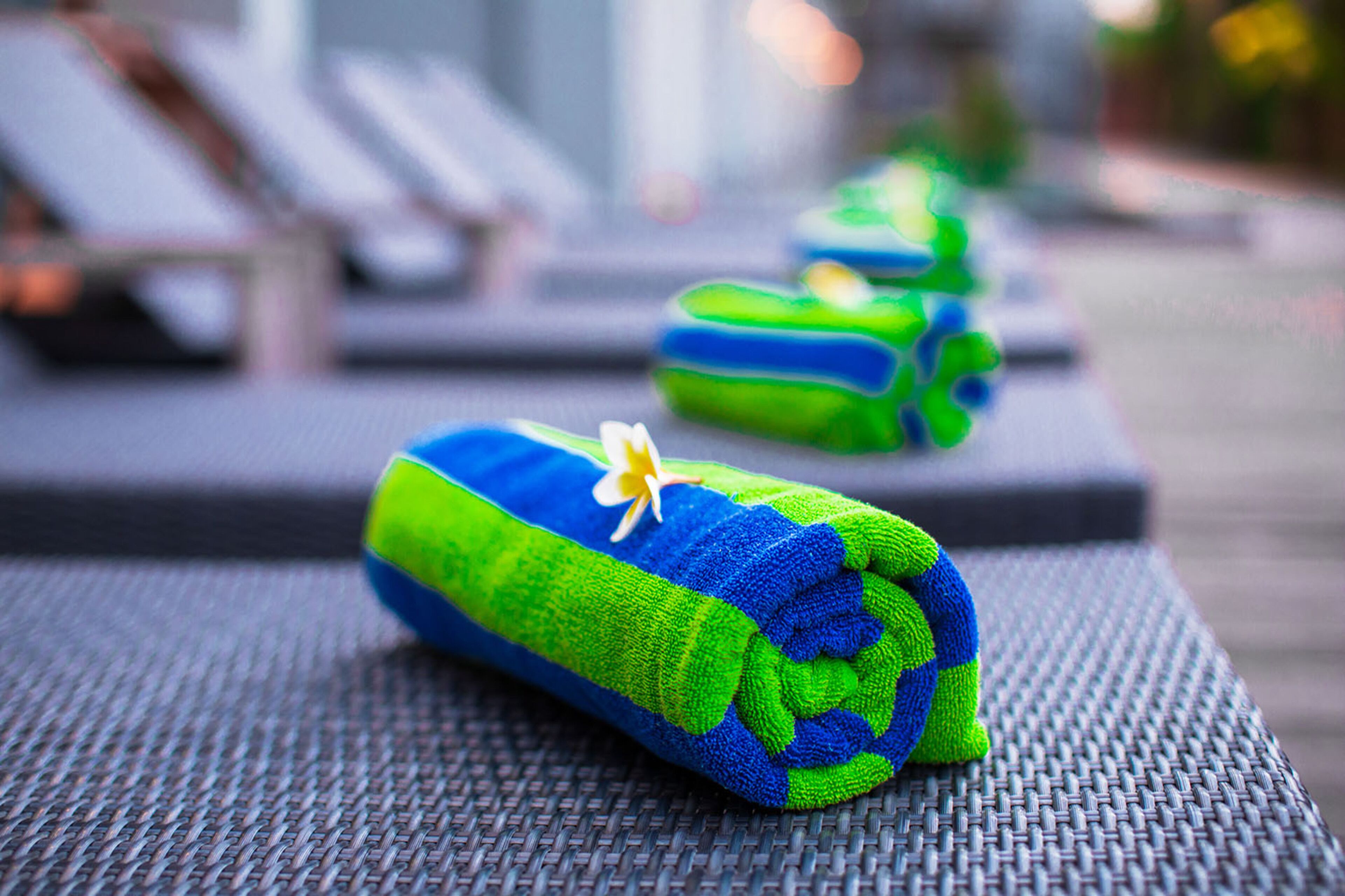 A rolled towel with a yellow flower on a textured surface, likely a poolside area with lounge chairs in the background.