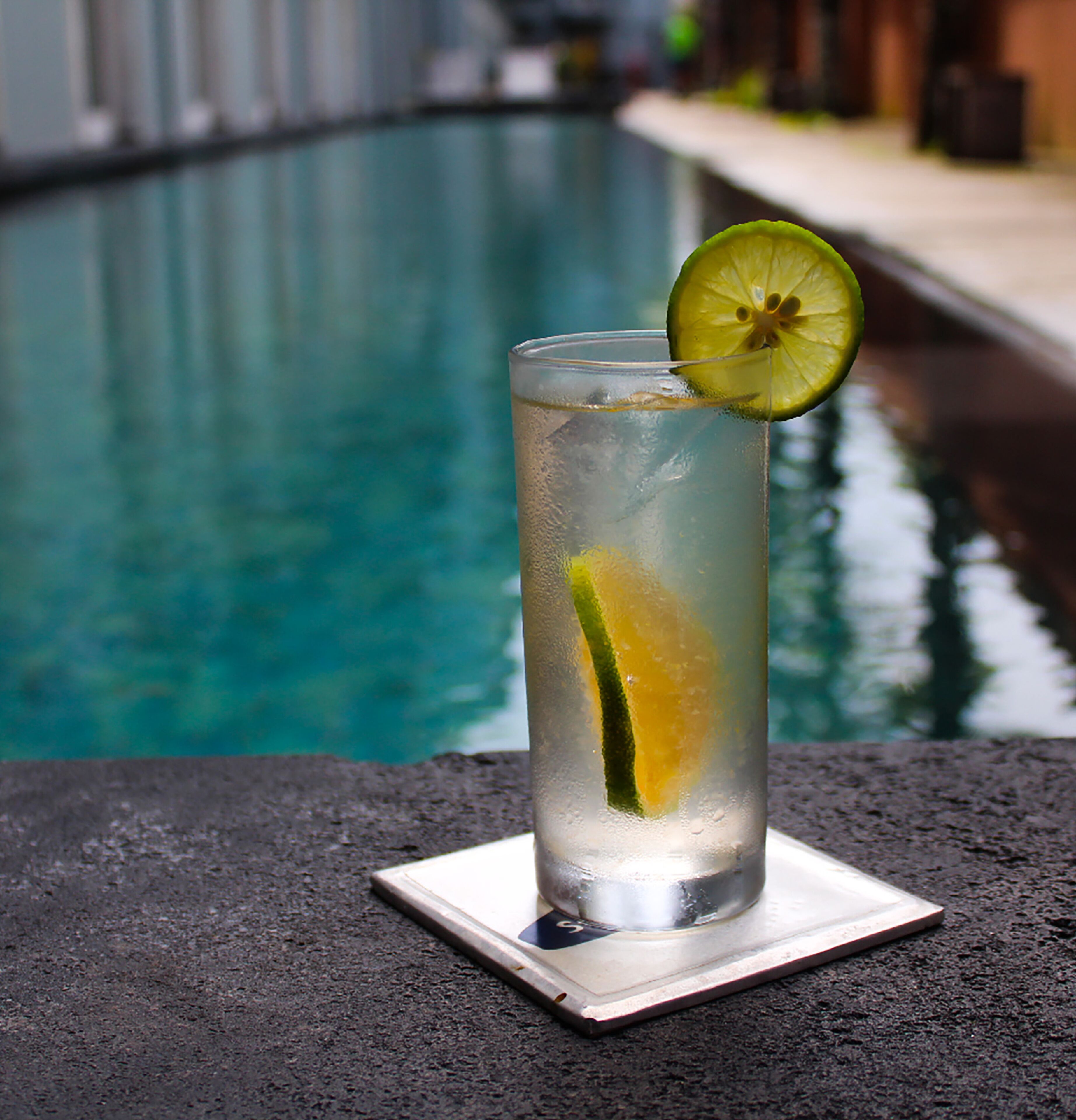 A tall glass with a lime slice garnish, placed on a coaster by a pool.