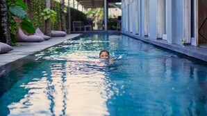 A swimming pool with a person in the water, surrounded by a covered patio area with seating and greenery. - FRii Bali Echo Beach (Canggu)