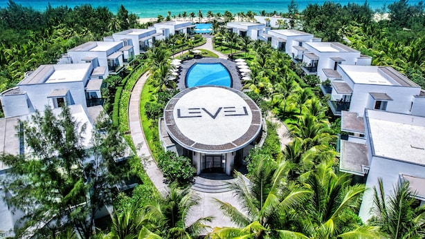 A resort with a central pool, surrounded by modern villas, palm trees, and a view of the ocean.