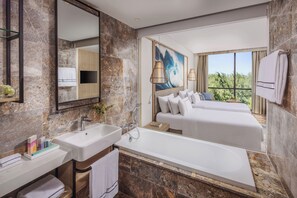 Un baño de hotel moderno con una bañera grande, una cama doble con ropa blanca y un espejo amplio.