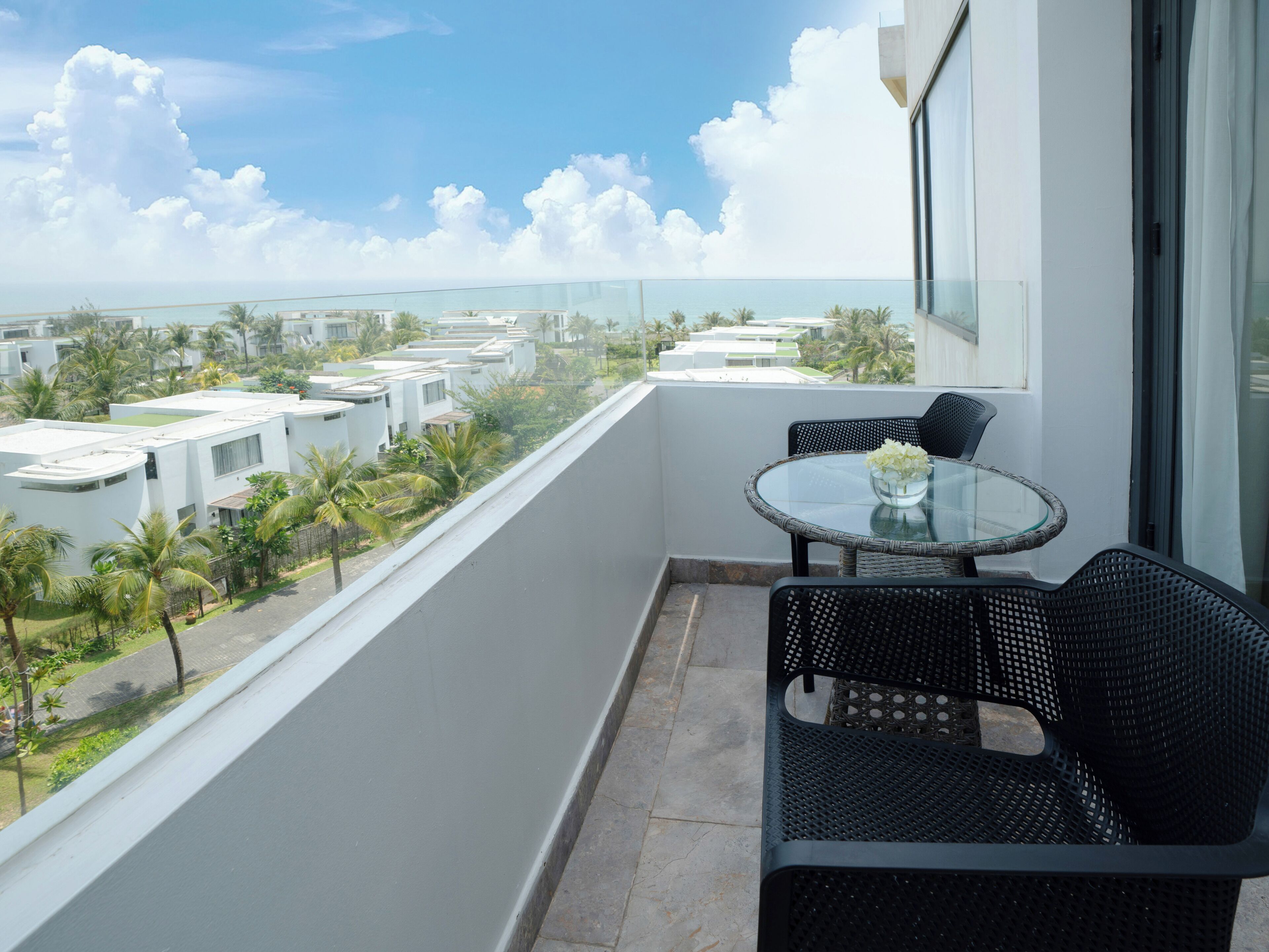 A balcony with a glass table and black wicker chairs, overlooking a residential area with palm trees and modern buildings.