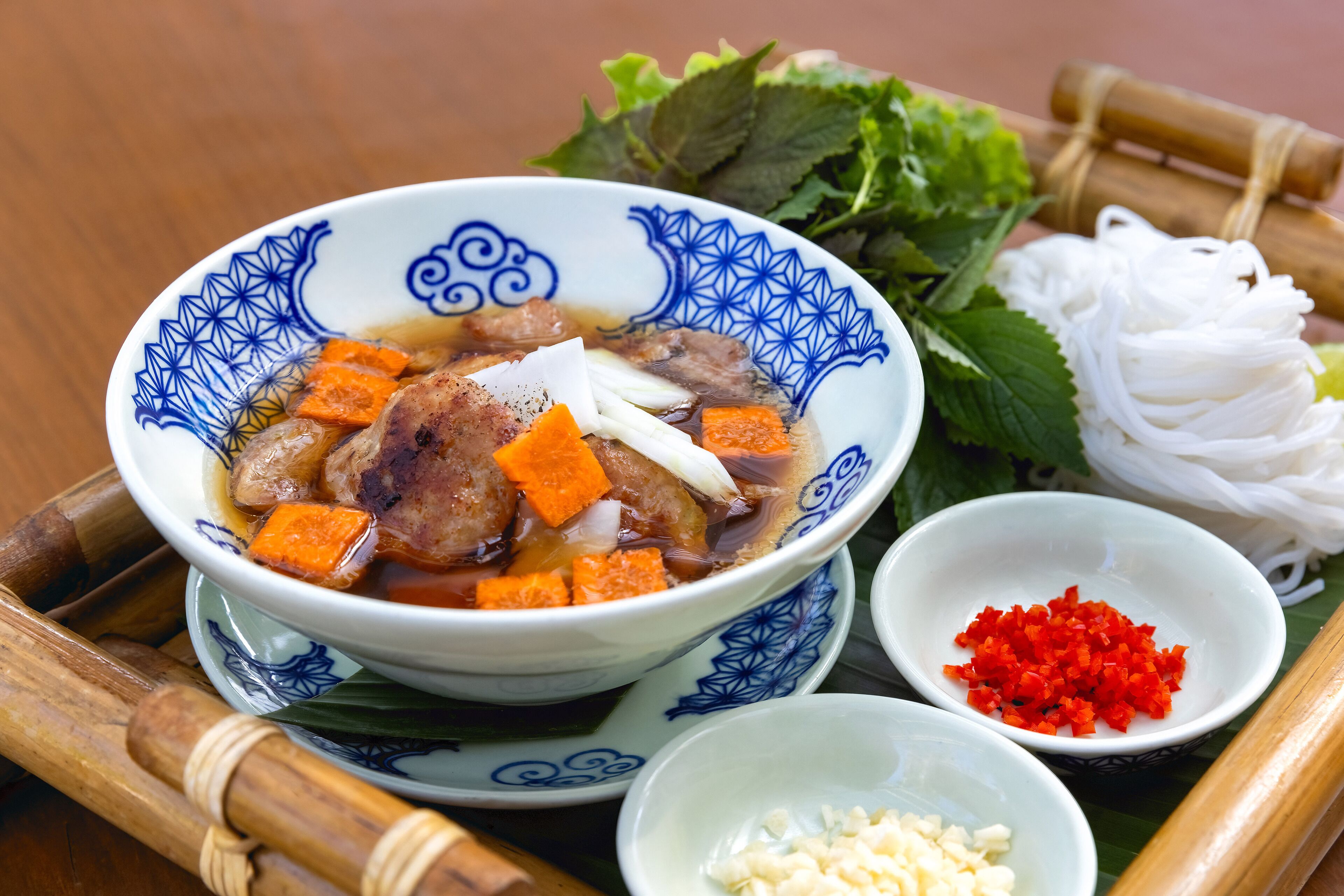 A bowl of soup with meat and vegetables, accompanied by fresh herbs and sliced noodles on a bamboo tray.