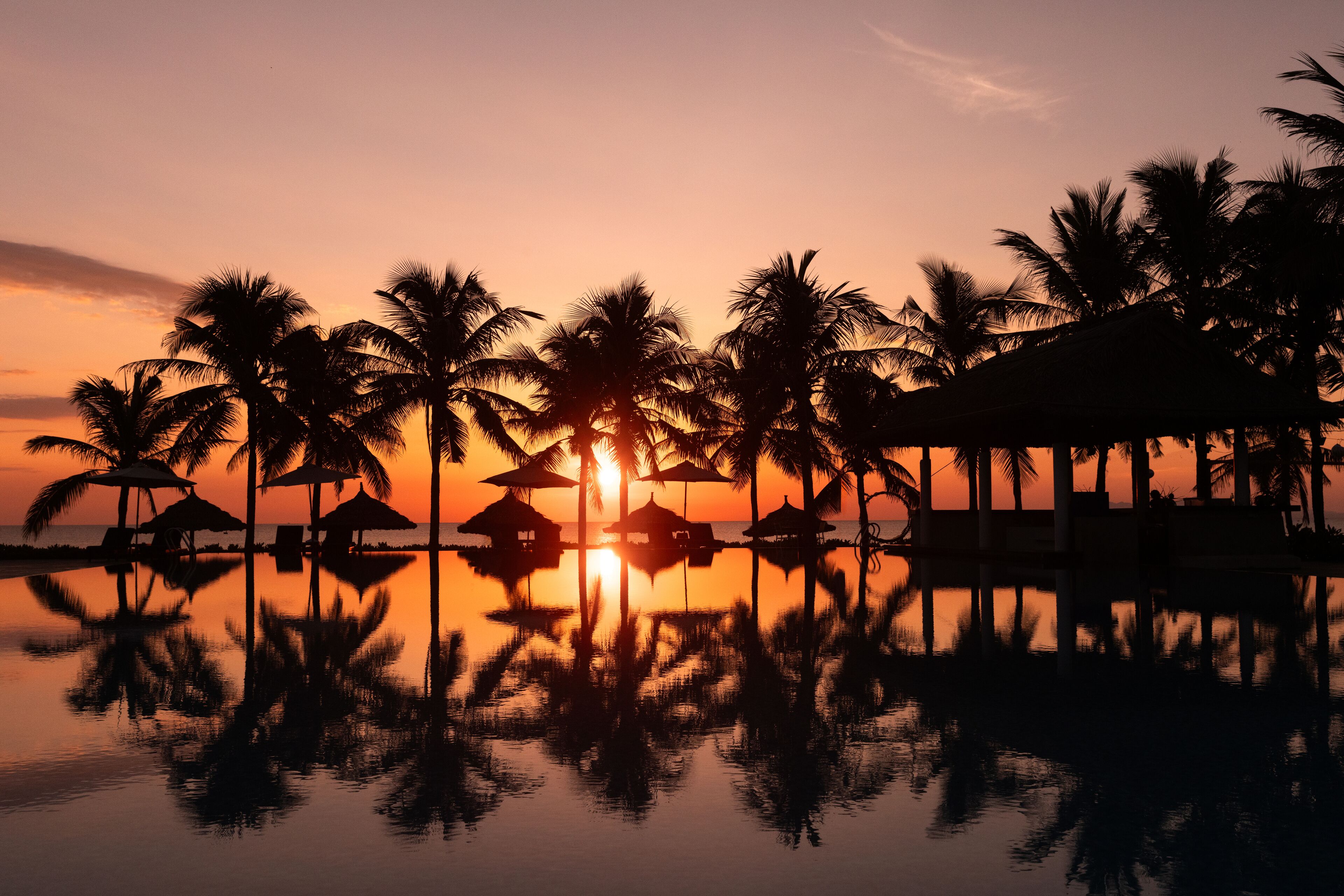Un atardecer sobre una masa de agua tranquila, con palmeras y bungalows sobre el agua.