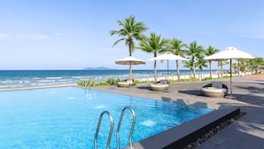 A swimming pool with clear blue water, surrounded by lounge chairs and umbrellas, with a beach and ocean view in the background.