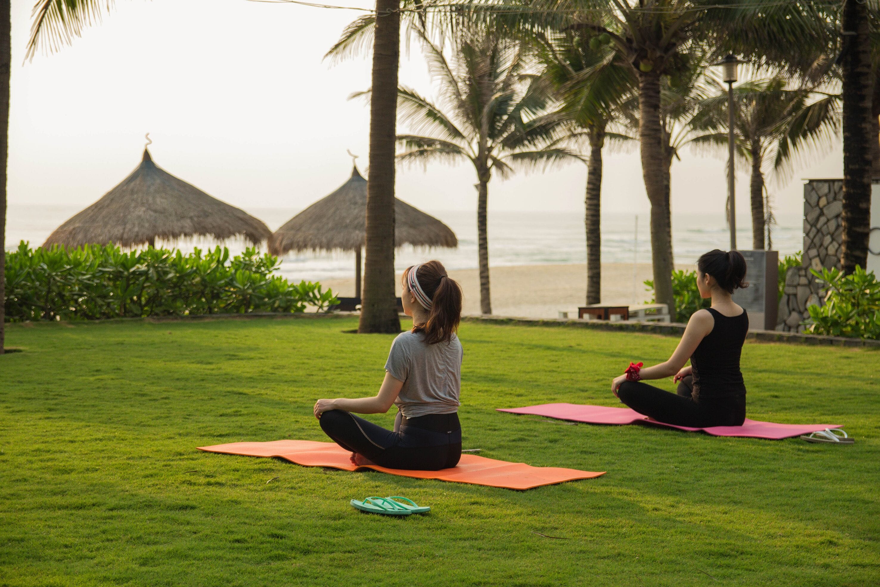 Two people meditating on grass mats with a beach and palm trees in the background.