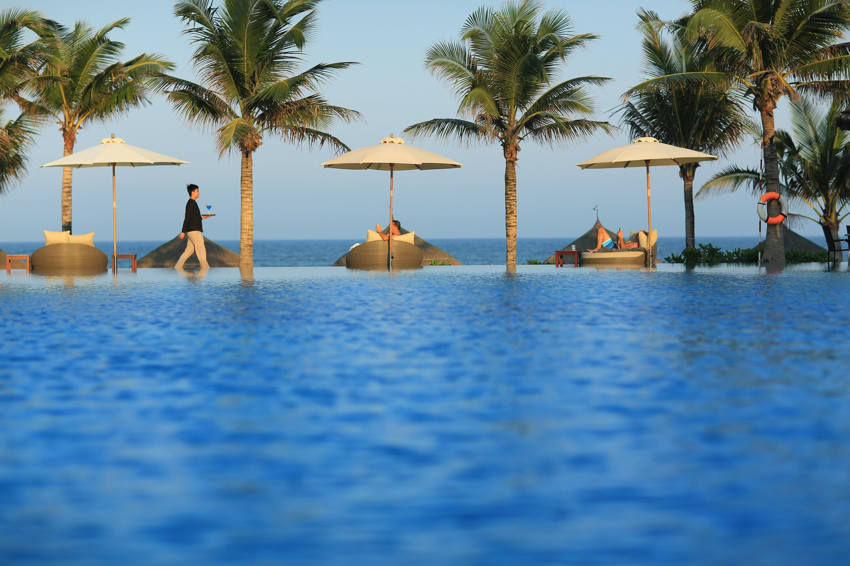 A pool area with palm trees and umbrellas, a lifebuoy on a palm tree, and a person walking.