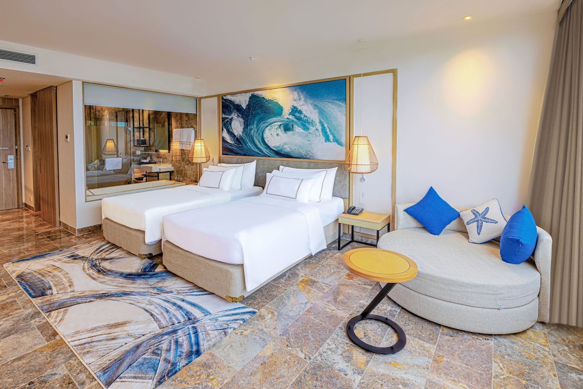A hotel room with two beds, a sofa, a small table, a large mirror, and a painting of a wave.