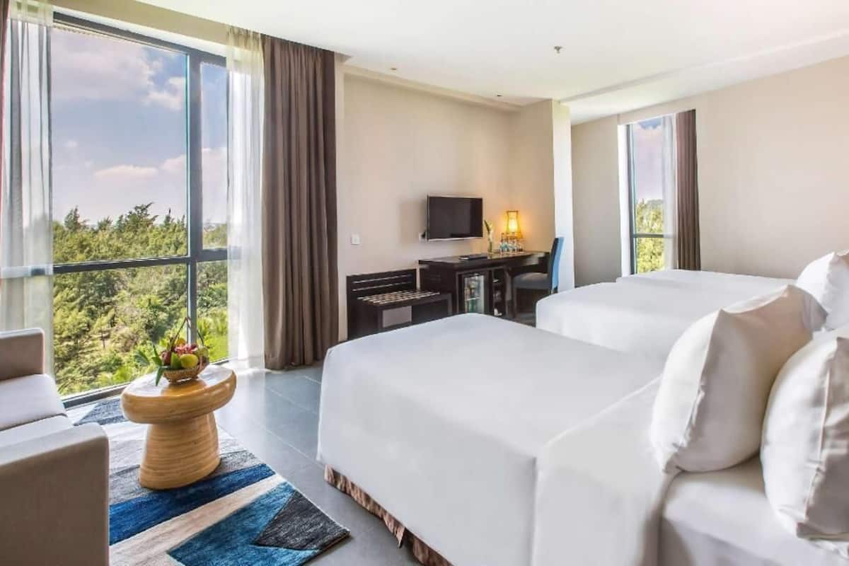 Ett hotellrum med två sängar, ett skrivbord, en tv och ett stort fönster med utsikt över grönområden.
