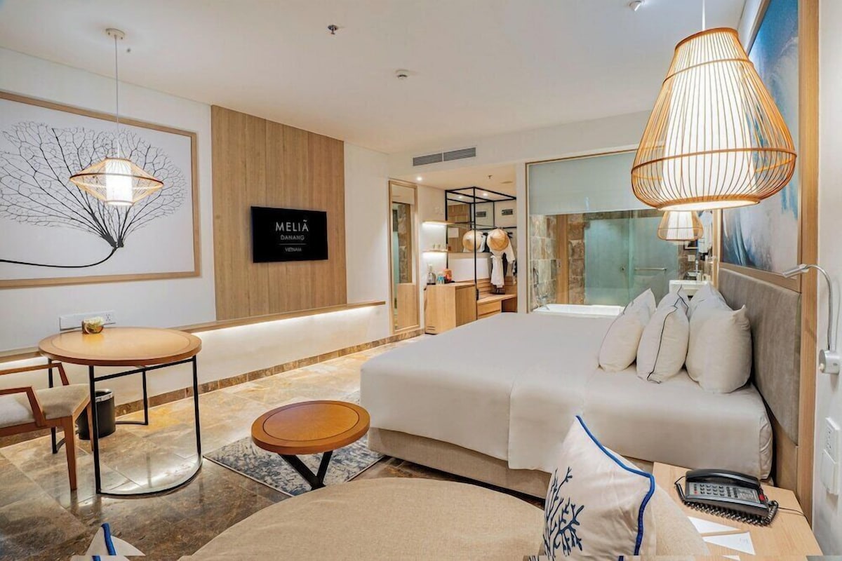 A modern hotel room with a large bed, a desk, a chair, a round table, and a wall-mounted TV.