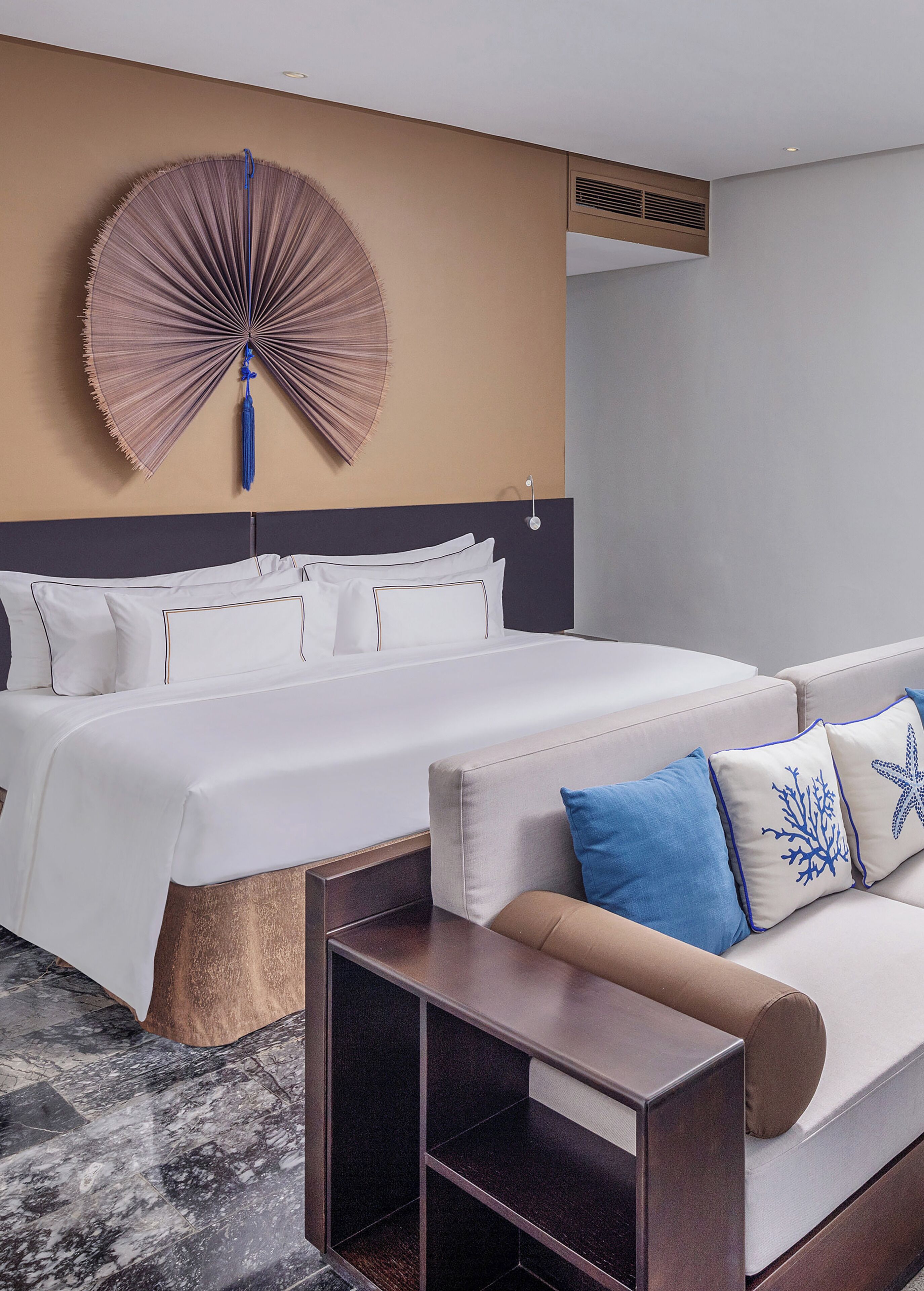 A hotel room with a bed, a sofa, a wooden side table, and a large wall fan.