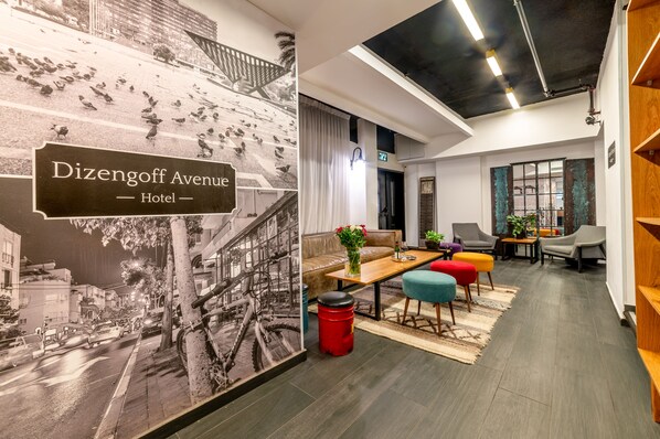 Un hall de hotel con un mural grande en blanco y negro de un paisaje urbano, un sofá, otomanas coloridas, una mesa de centro de madera y un bote de basura rojo.