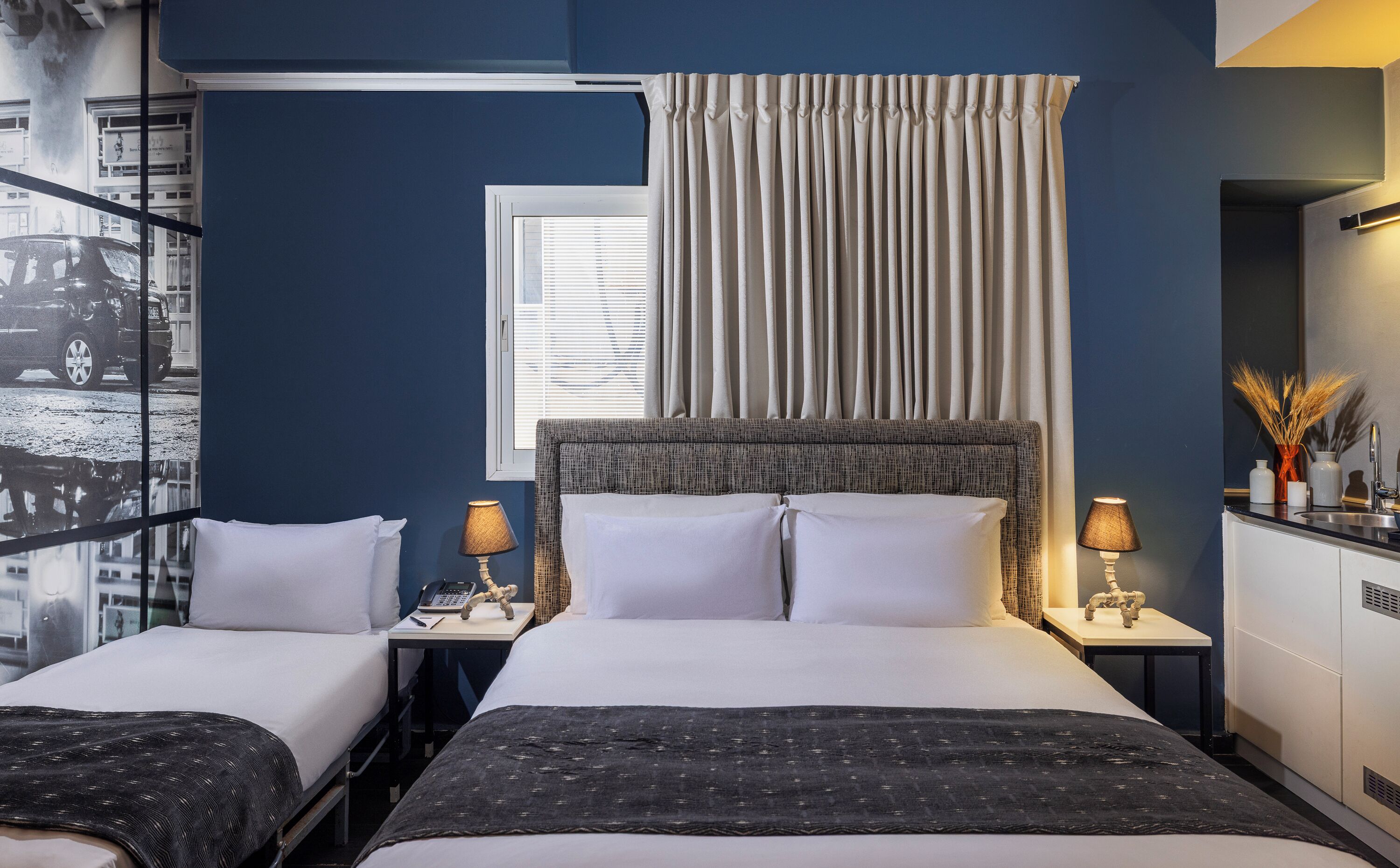 Une chambre d’hôtel avec deux lits, une fenêtre avec des stores, une tête de lit, des tables de chevet avec des lampes, un miroir et une vue sur la rue depuis la fenêtre.