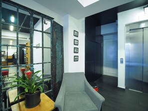 Un espacio de oficina moderno con área de recepción, un sofá gris, una mesa de madera con planta en maceta y un ascensor.
