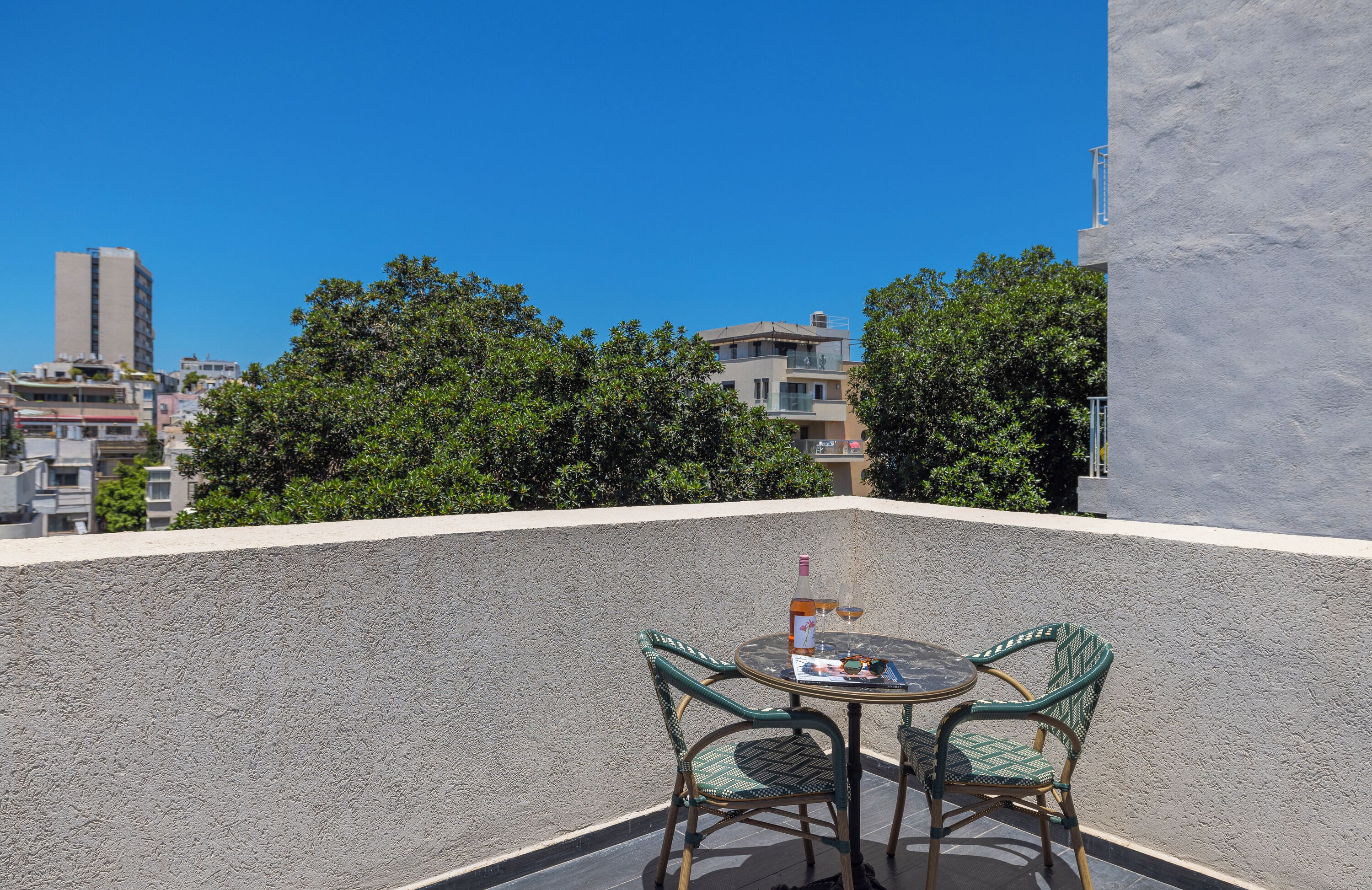 A rooftop terrace with a table and chairs, a bottle of wine, and a view of city buildings and trees.