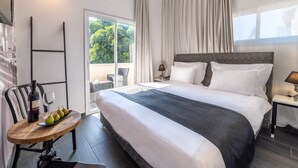 Quarto de hotel com uma cama grande, uma mesa pequena com uma garrafa e copos, e vista para o verde lá fora.