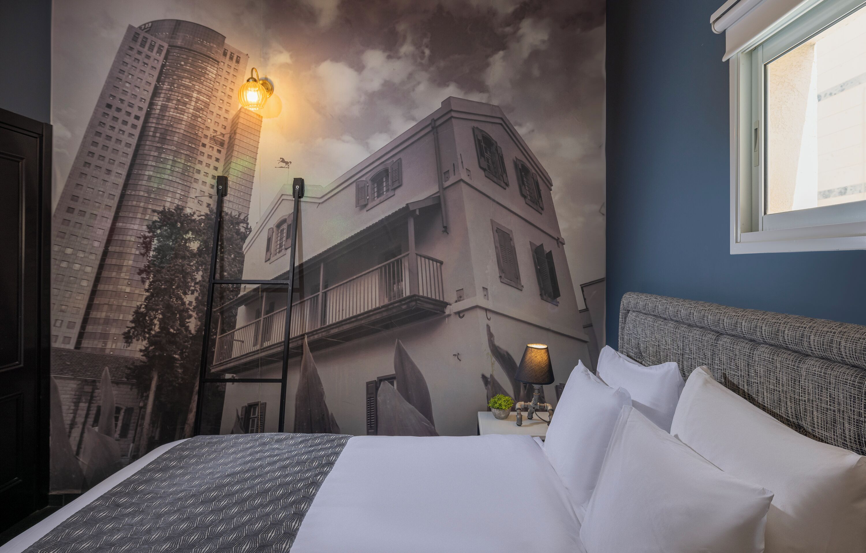 A hotel room with a bed, a nightstand with a lamp, a wall mural of a building, and a window.