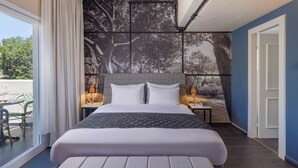 Quarto de hotel moderno com uma cama grande, vista para árvores pela janela e uma mesinha com abajur.
