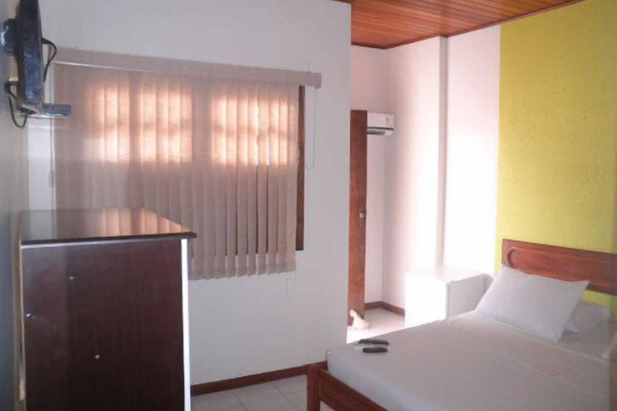A hotel room with a wooden ceiling, a bed with white bedding, a desk with a computer, and a window with vertical blinds.