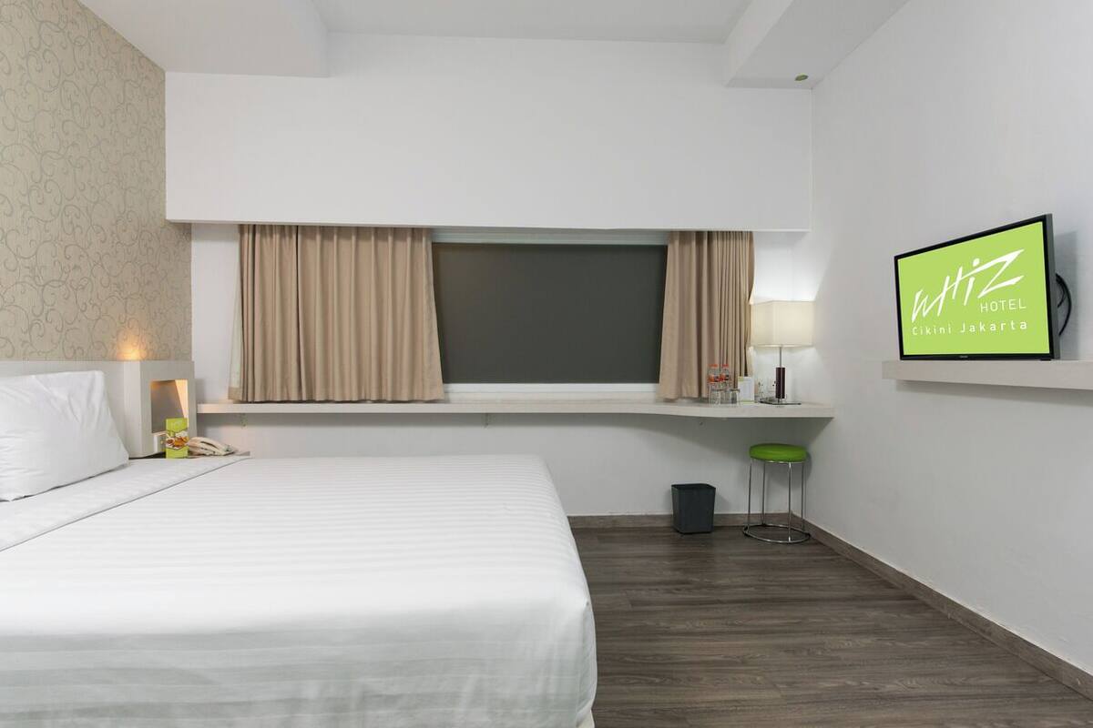 A hotel room with a bed, a TV, a window with curtains, a small table with a lamp, and a trash bin.