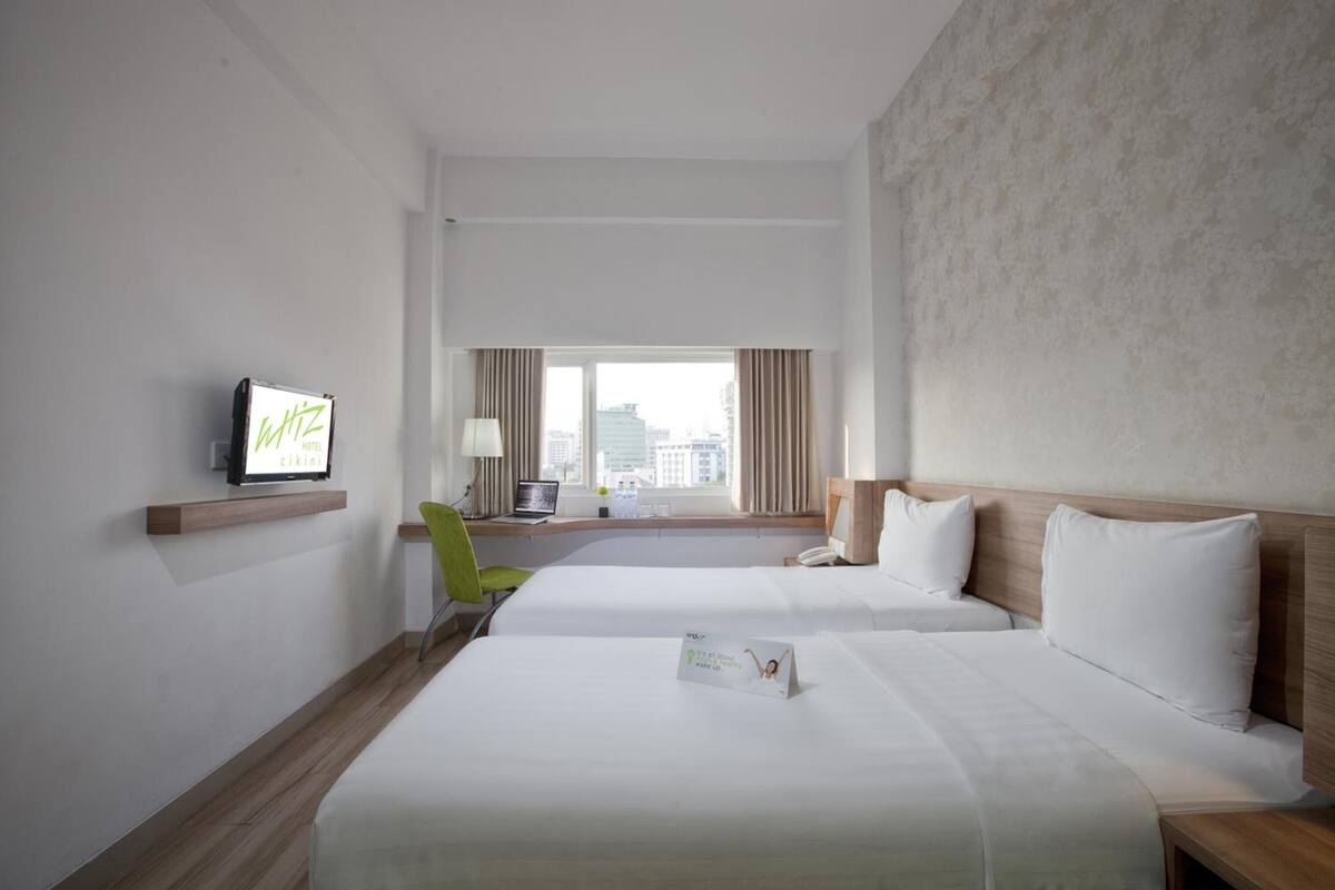 A hotel room with two beds, a desk with a laptop, a television, and a view of the city.