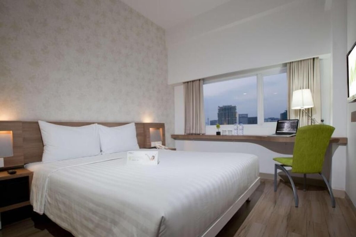 Habitación de hotel con una cama grande, un escritorio con una silla, una ventana con vistas a edificios y una lámpara en el escritorio.