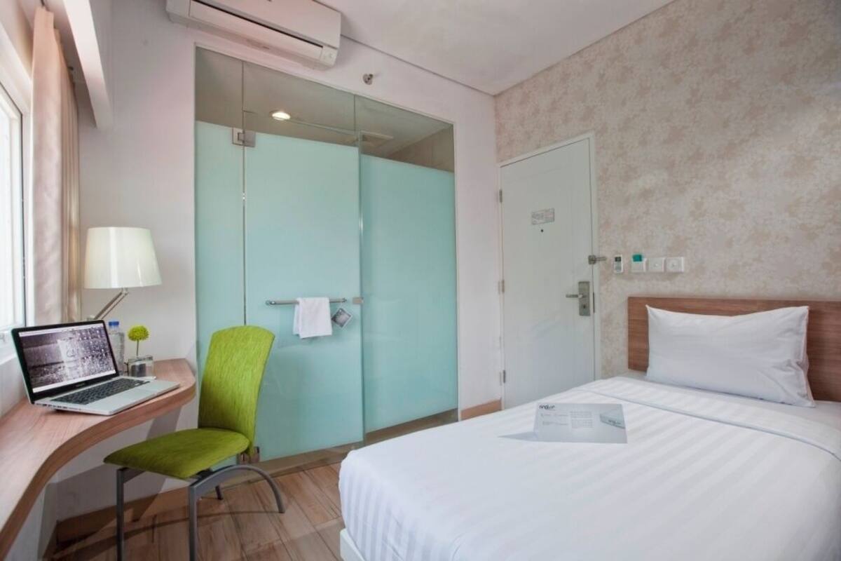A hotel room with a bed, a desk with a laptop, a green chair, a glass door closet, and a wall-mounted air conditioner.