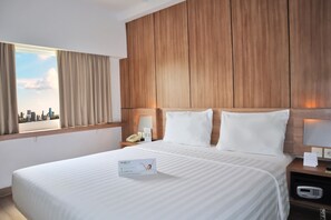 Habitación de hotel con una cama grande, cabecera de madera, mesita de noche con teléfono y mesita auxiliar con lámpara, además de una ventana con cortinas.
