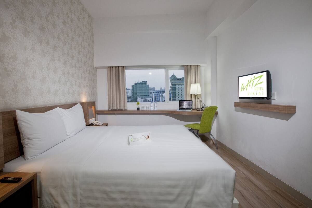 A hotel room with a large bed, a desk with a laptop, a television, and a view of the cityscape through the window.