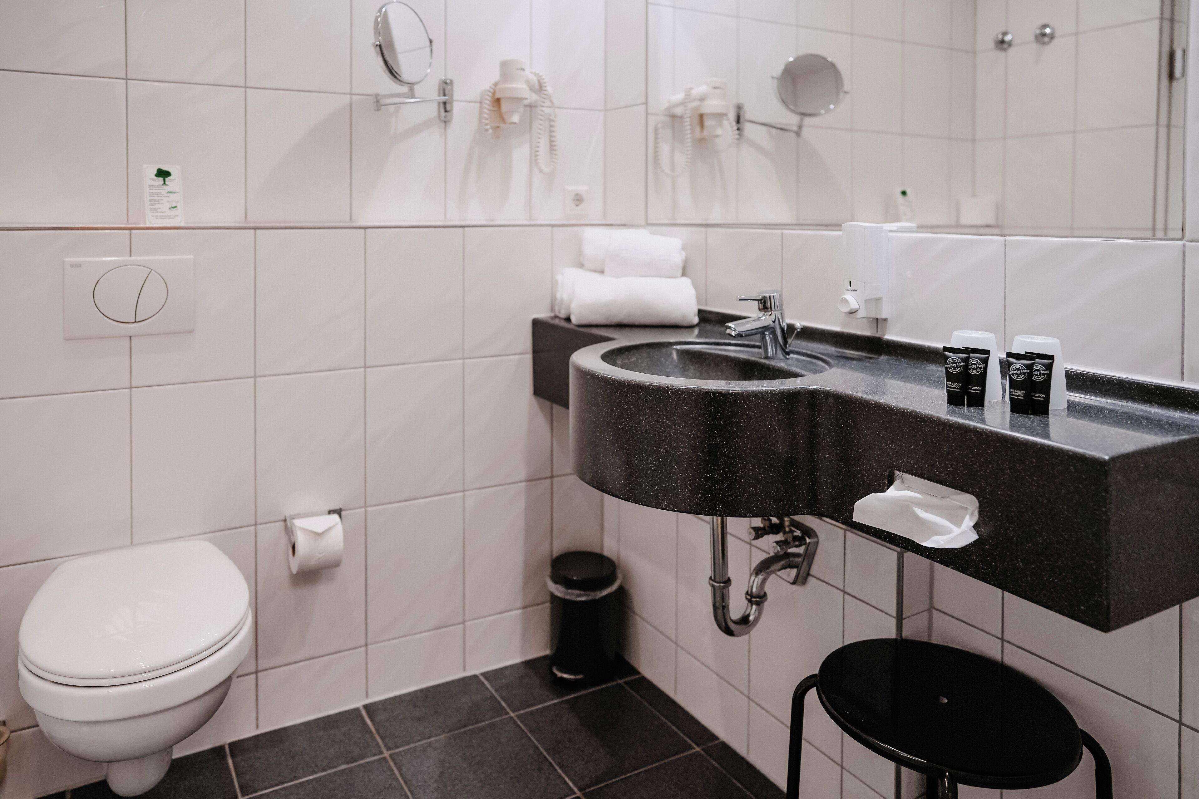 A modern bathroom with a black sink, white tiles, a toilet, and a trash can.