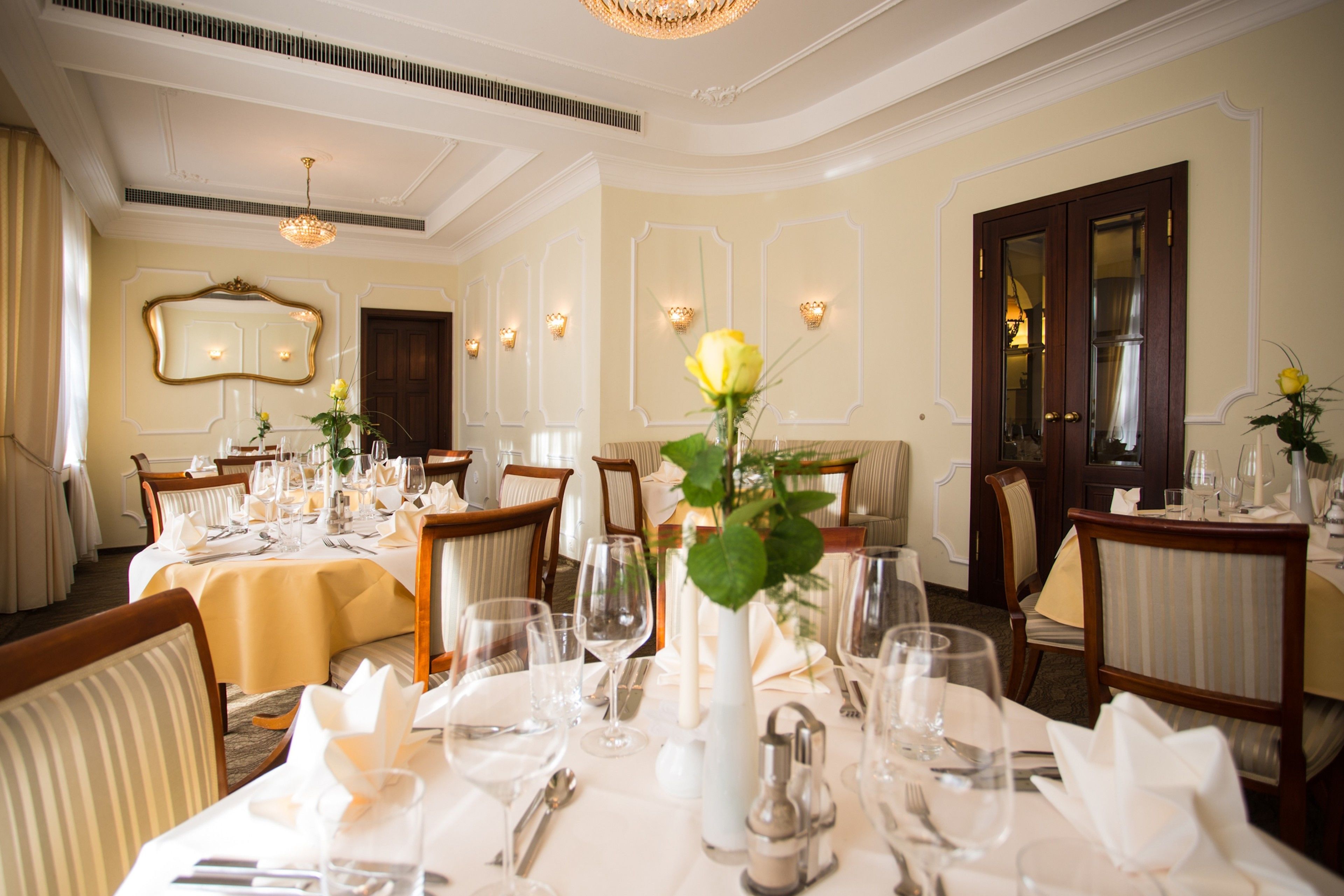 Uma sala de jantar formal com mesas preparadas para o serviço, luminárias elegantes e um espelho grande na parede.
